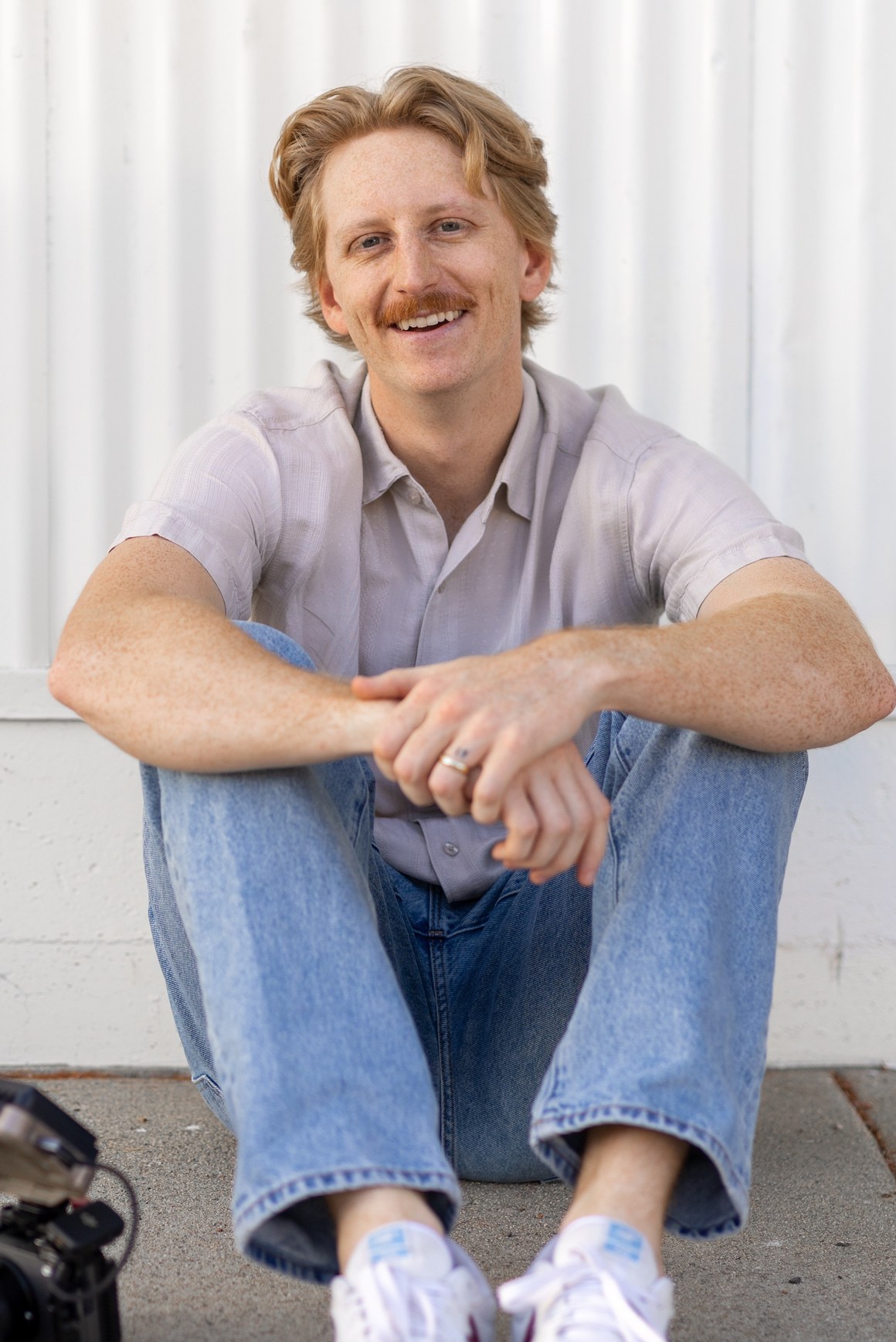 Daniel Lindeen sitting against a white corrugated wall with his camera beside him