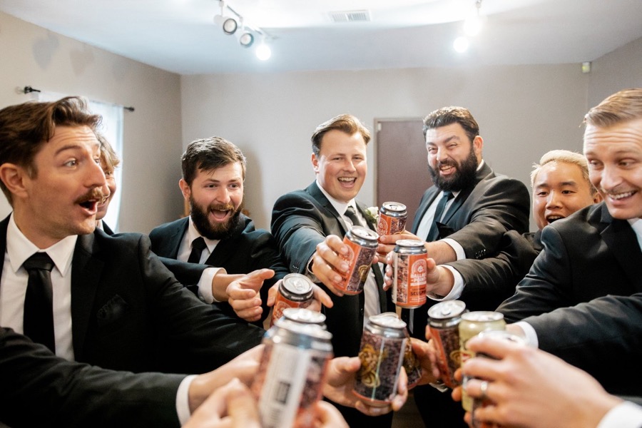 Groomsmen in black suits knocking cans together in a tight circle