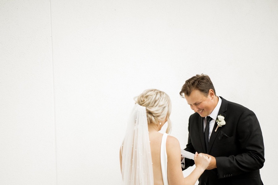 A groom looking down at his bride's hands against a bare white wall