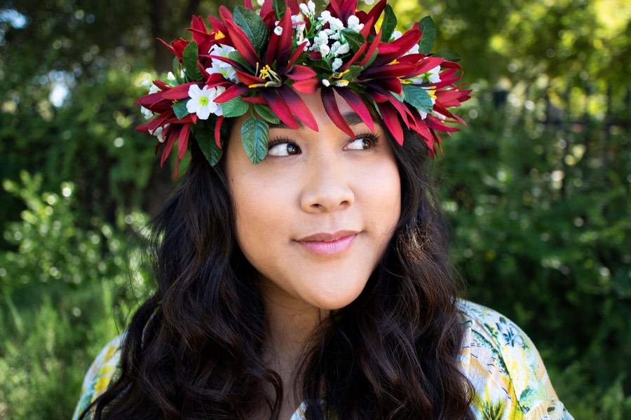 A woman in a red and white flower crown looking off camera against dense green foliage