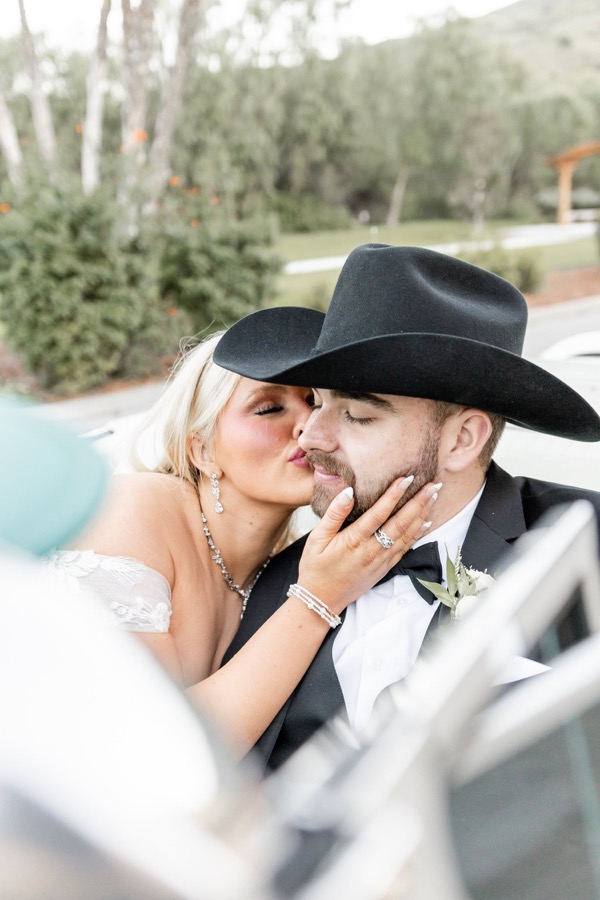 A bride kissing her groom's cheek, seen through a car window, his black cowboy hat tipped forward
