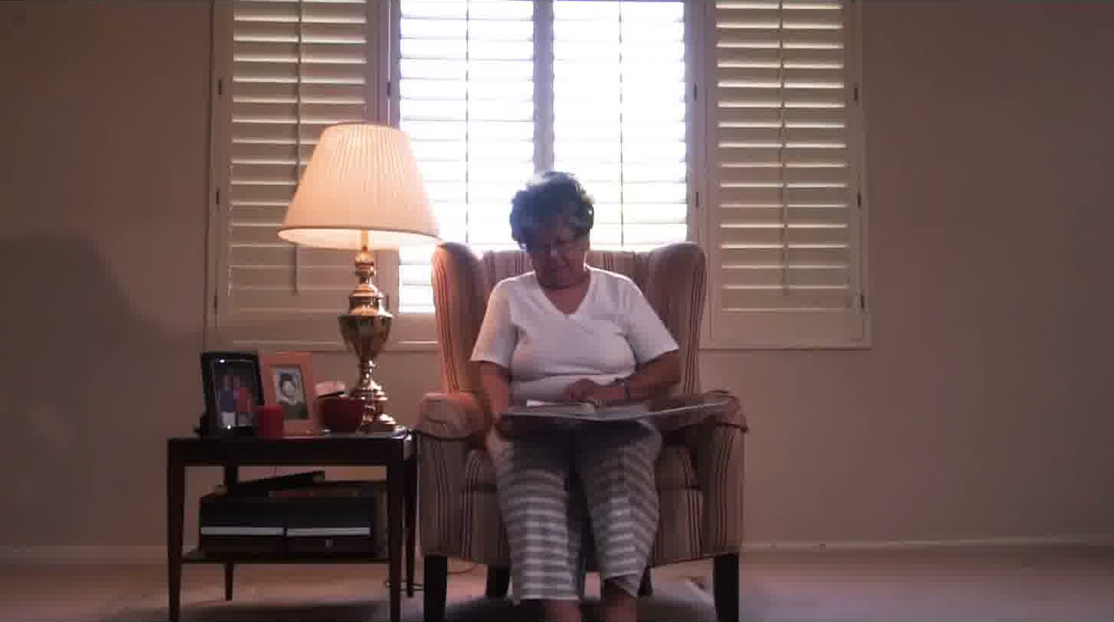 An elderly woman looking through a photo album in a wingback chair, lit by a window and a lamp